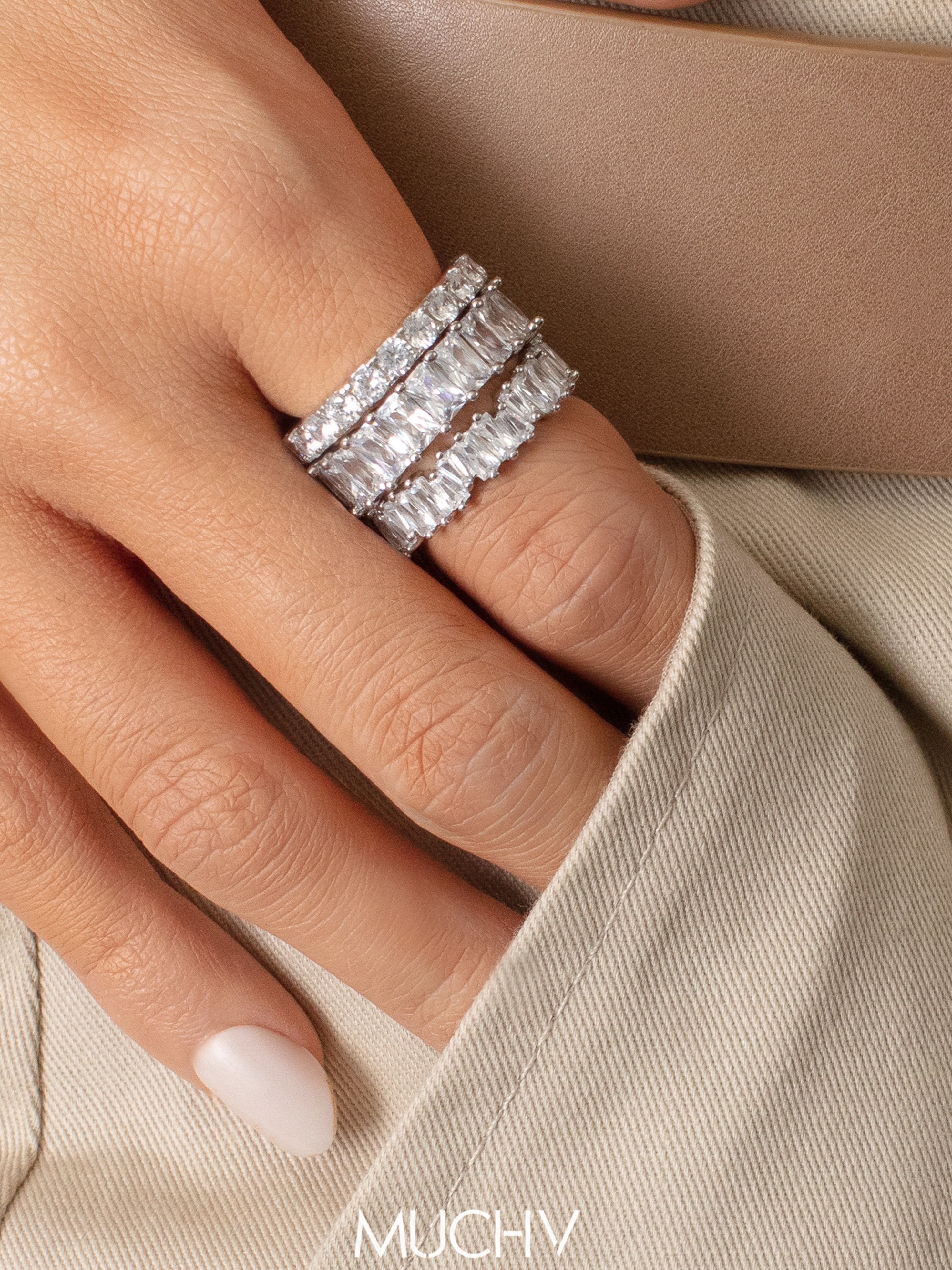 Hand with short pink nails wearing three sterling silver rings stacked together. Each ring band has different types of stones that range from round-shaped to emerald-shaped.