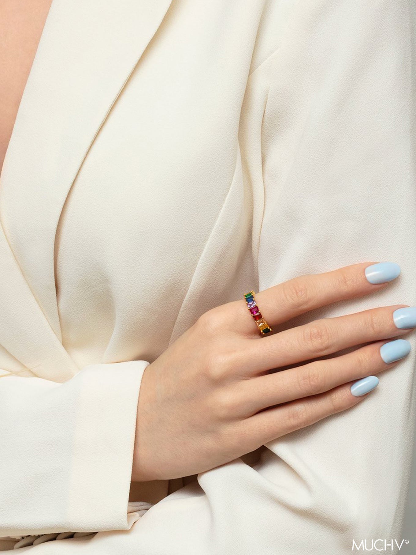 Model with pastel blue nails wearing a gold stacking ring with colourful rainbow stones.