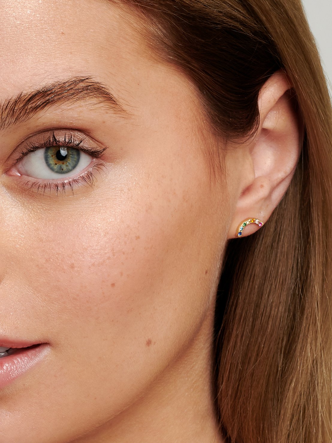 Teenage model wearing gold rainbow shaped stud earrings with colourful stones.