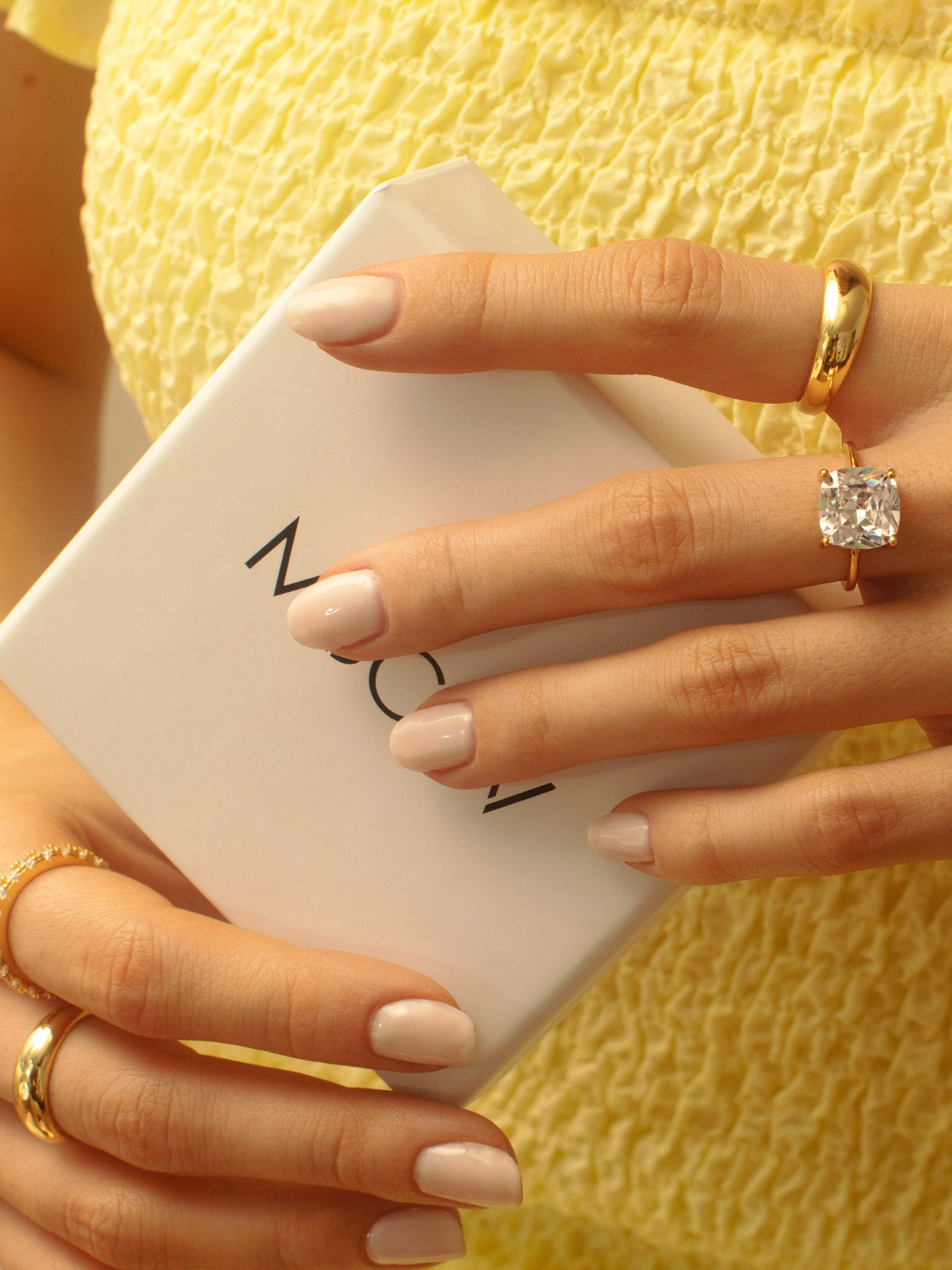 Model wearing a gold ring with a square cubic zirconia stone. Paired together with a gold dome ring and short nude nails.